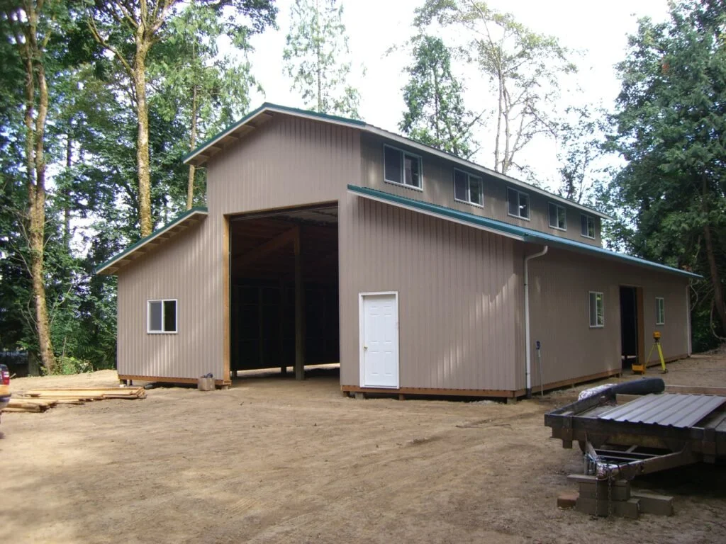 Newly constructed barn or large shed with beige siding, multiple small windows, and a sliding door, set in a wooded area with dirt ground.