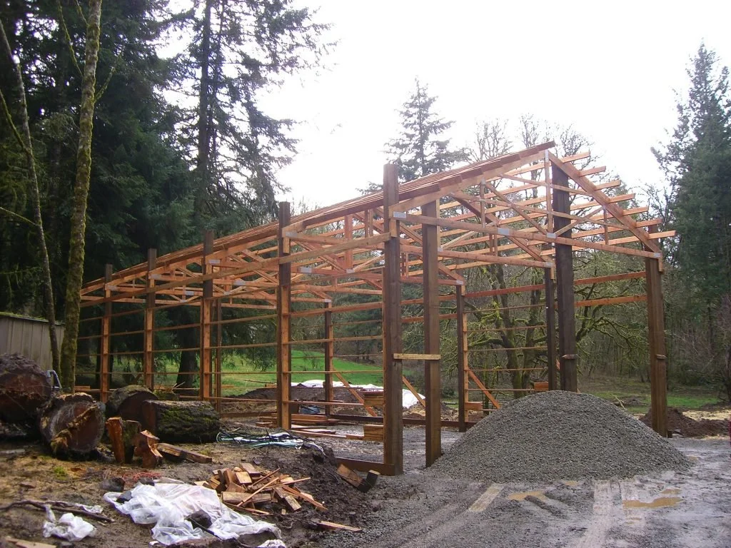 A wooden structure under construction in a forested area, with a pile of gravel and logs nearby.