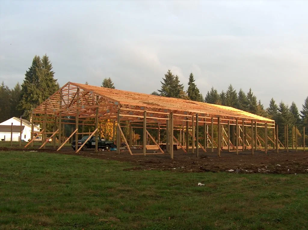 A wooden frame structure for a building under construction in a grassy area, with trees and a house in the background.