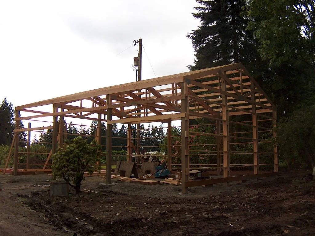Wooden frame of a building under construction on a dirt lot with trees and an electrical pole in the background.