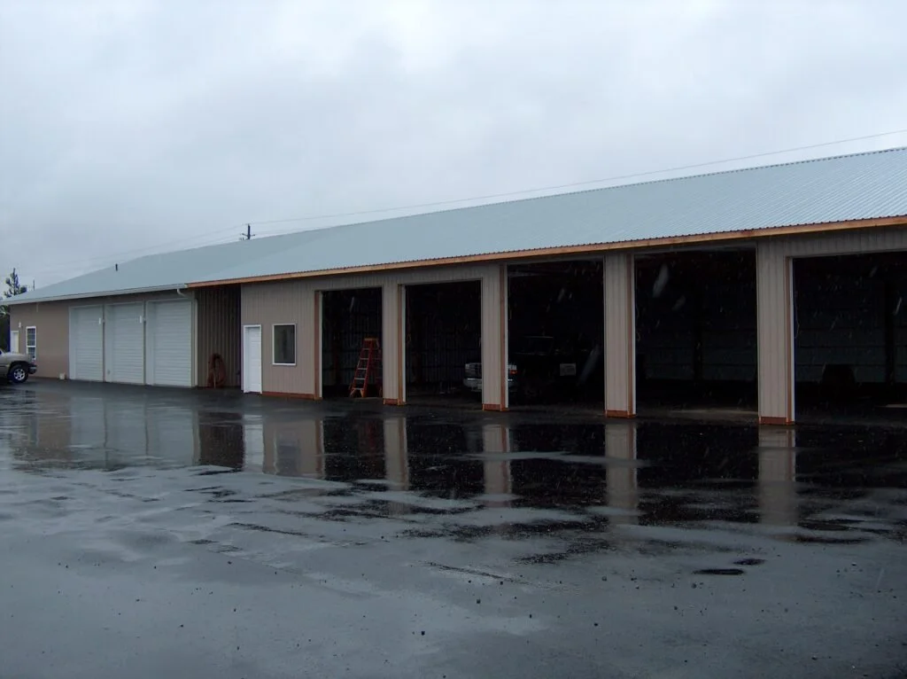 A large building with multiple garage doors and open bays, on a wet pavement on a cloudy day.