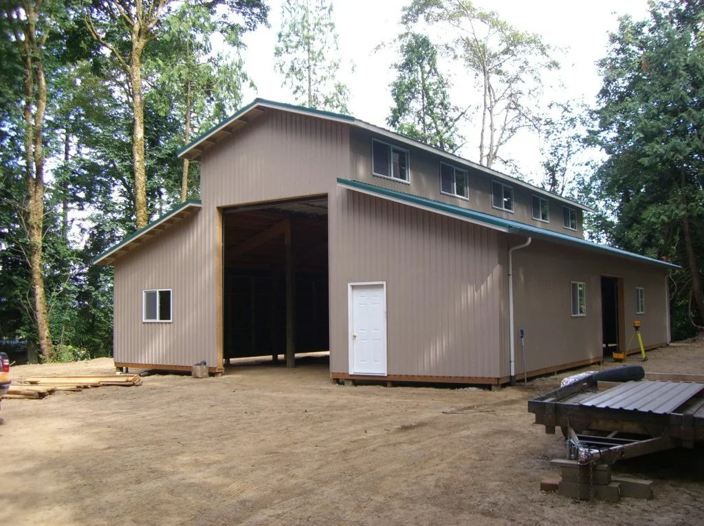 A two-story barn under construction in a wooded area with dirt ground, with windows, a white door, and building materials nearby.