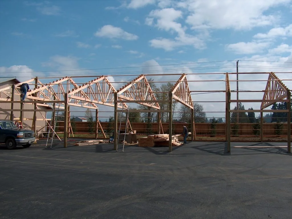 Construction workers building a wooden framework for a structure, with scaffolding and ladders, near a parking lot under a partly cloudy sky.