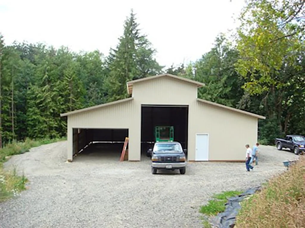 A large beige metal building with a sloped roof, two large garage doors, and a man and woman walking nearby with trees in the background.