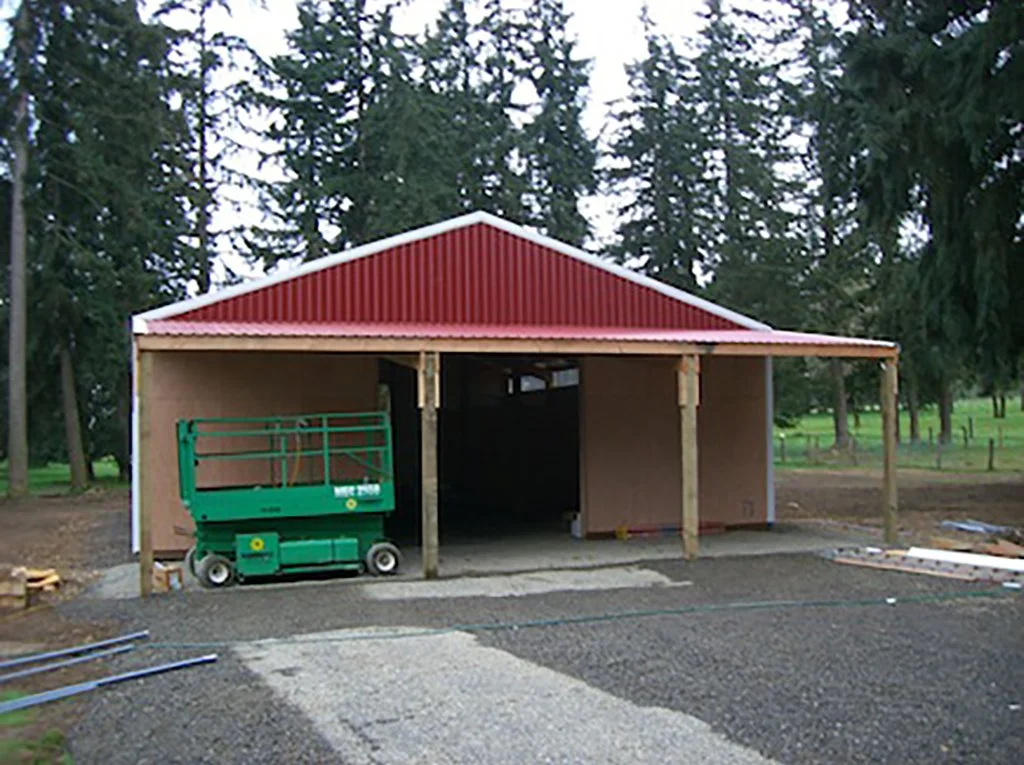 Construction site of a small open structure with a red metal roof and unfinished wooden walls, surrounded by trees, a green lift, and building materials.