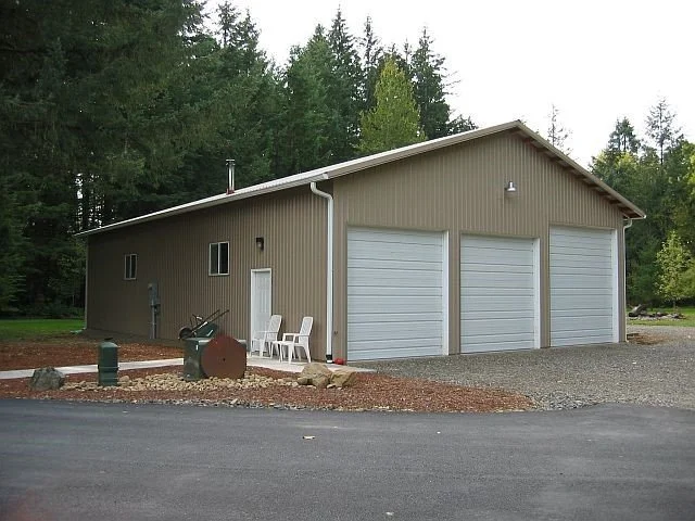 A beige metal garage with three white garage doors, situated in front of a wooded area, with a small gravel driveway and some outdoor chairs near the side.