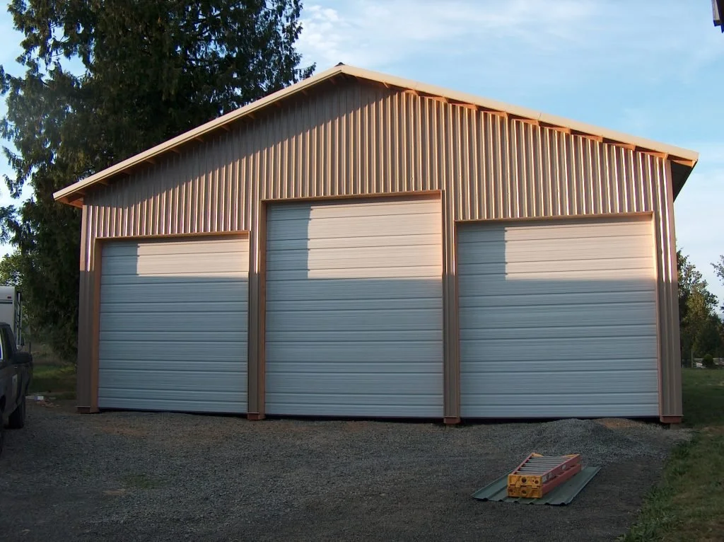 A metal garage with three large roll-up doors, situated on a gravel lot with tools and a vehicle nearby, surrounded by trees and under a partly cloudy sky.