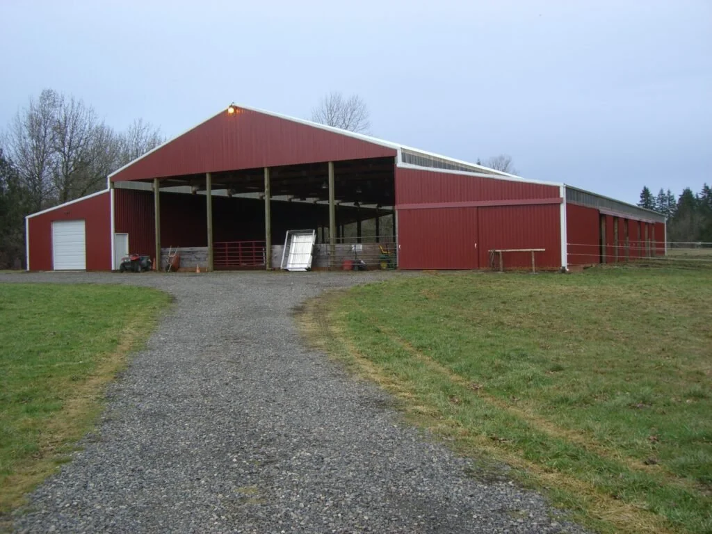 Red barn with open section and closed garage-style door, located on a gravel driveway surrounded by grass and trees.