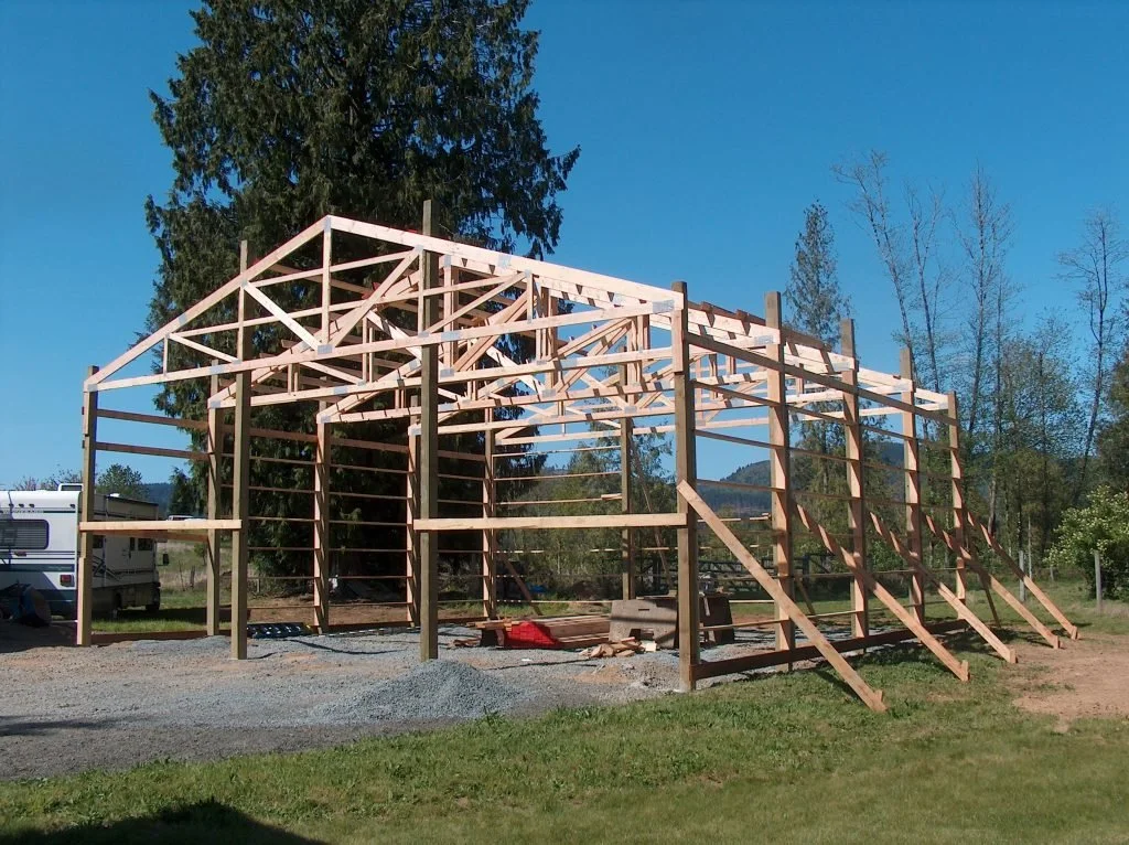 Construction of a wooden building frame outdoors on a clear day, with a blue sky, trees, and grass visible.