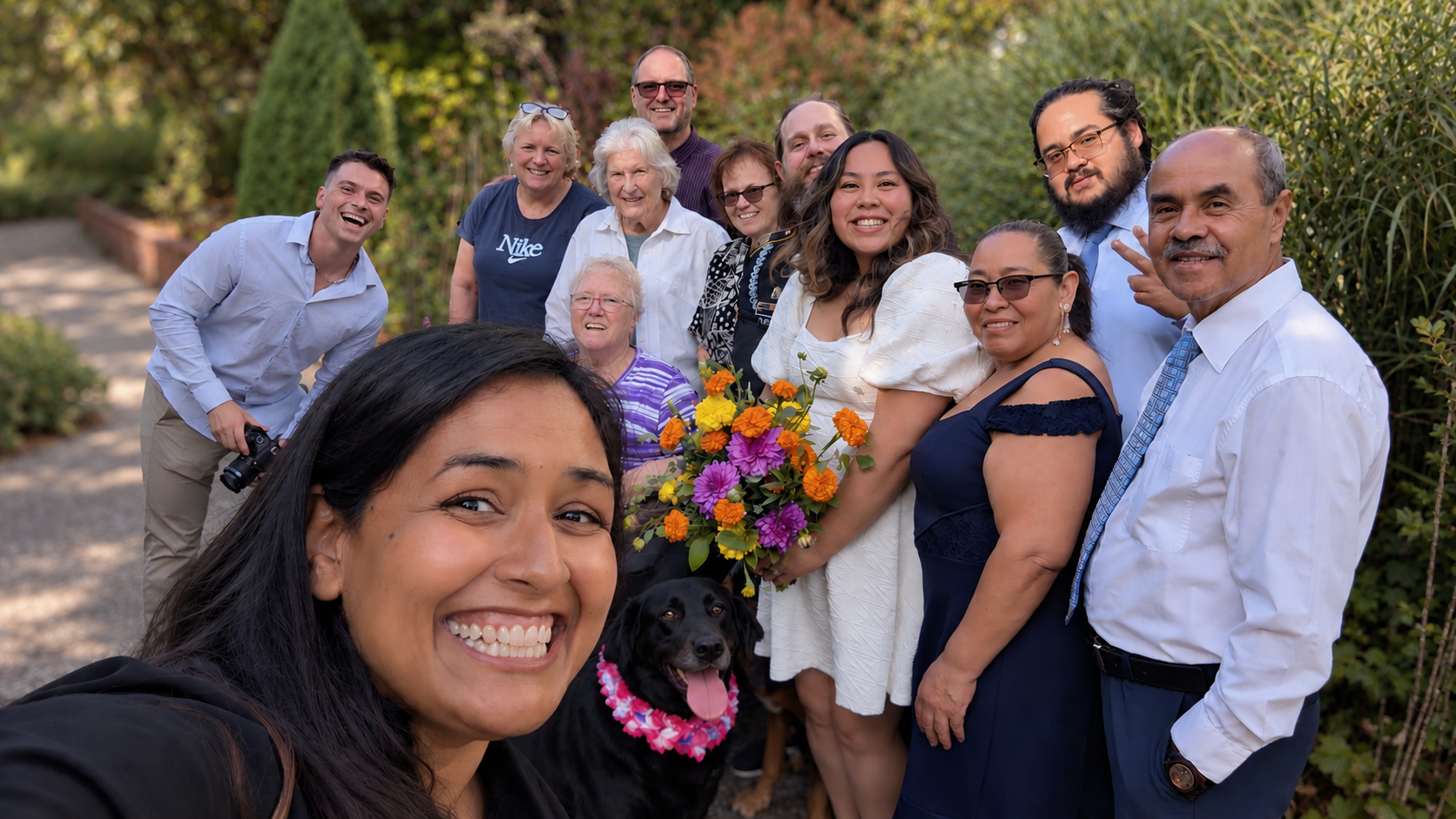 A group of diverse people smiling outdoors, taking a selfie with a woman in the front holding a bouquet of flowers and a black dog wearing a pink floral bandana.