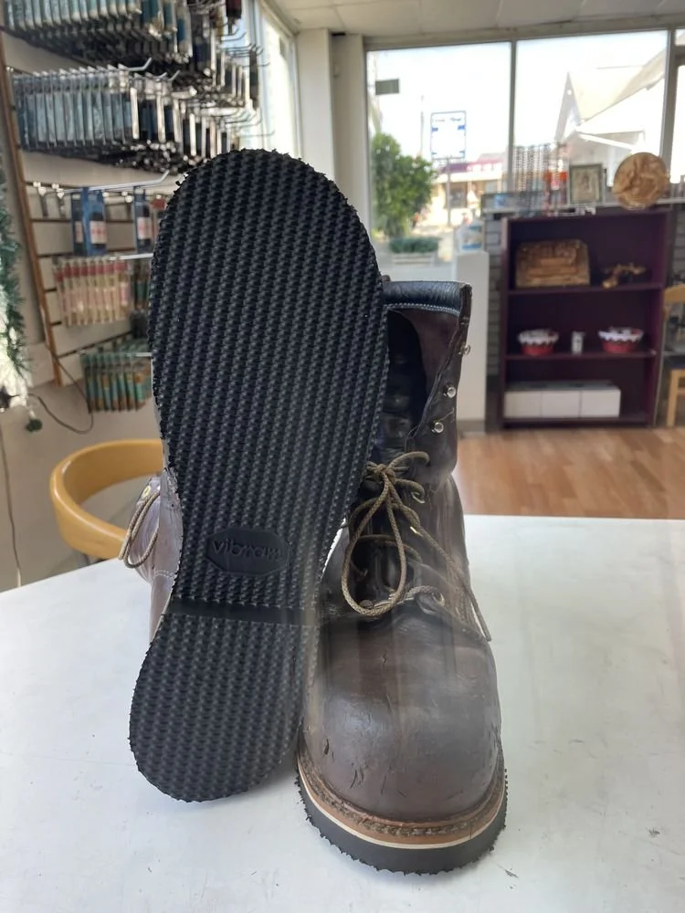 A pair of dark brown leather boots with laces, positioned on a white table in a store, with one boot's sole visible showing a textured black Vibram rubber bottom, inside a store with shelves, a window, and various items in the background.