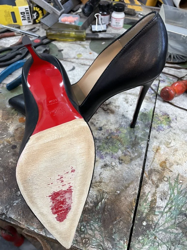 A pair of black high-heeled shoes, one showing the bottom with a red sole and some scuffs, placed on a cluttered work table with tools, paint bottles, and other craft supplies.