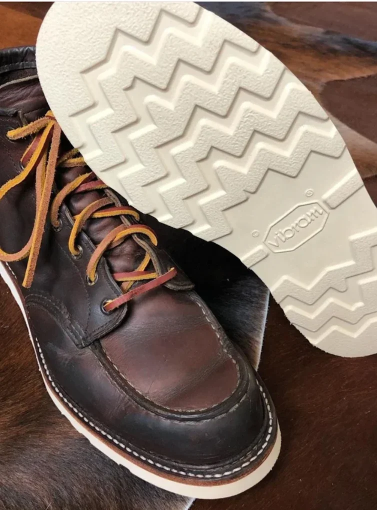 Close-up of a brown leather boot with yellow laces, showing the sole with a zigzag tread pattern and the brand name 'Vibram' embossed on it, placed on a wooden surface.
