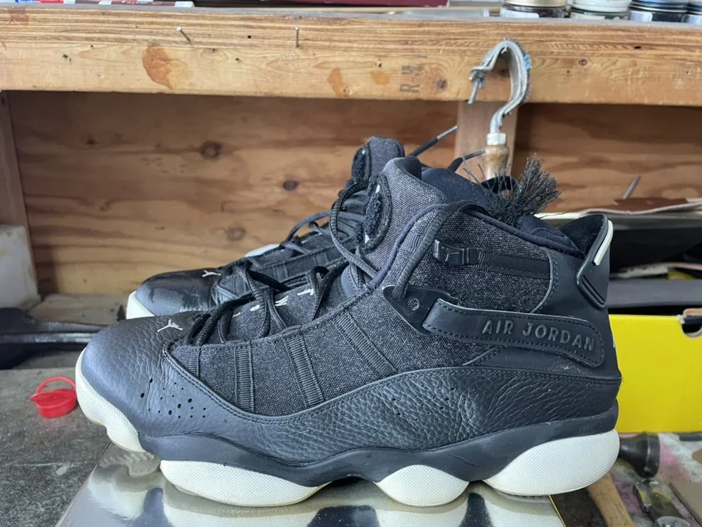 A pair of black Nike Air Jordan sneakers with white soles, placed on a reflective surface in a workshop setting.
