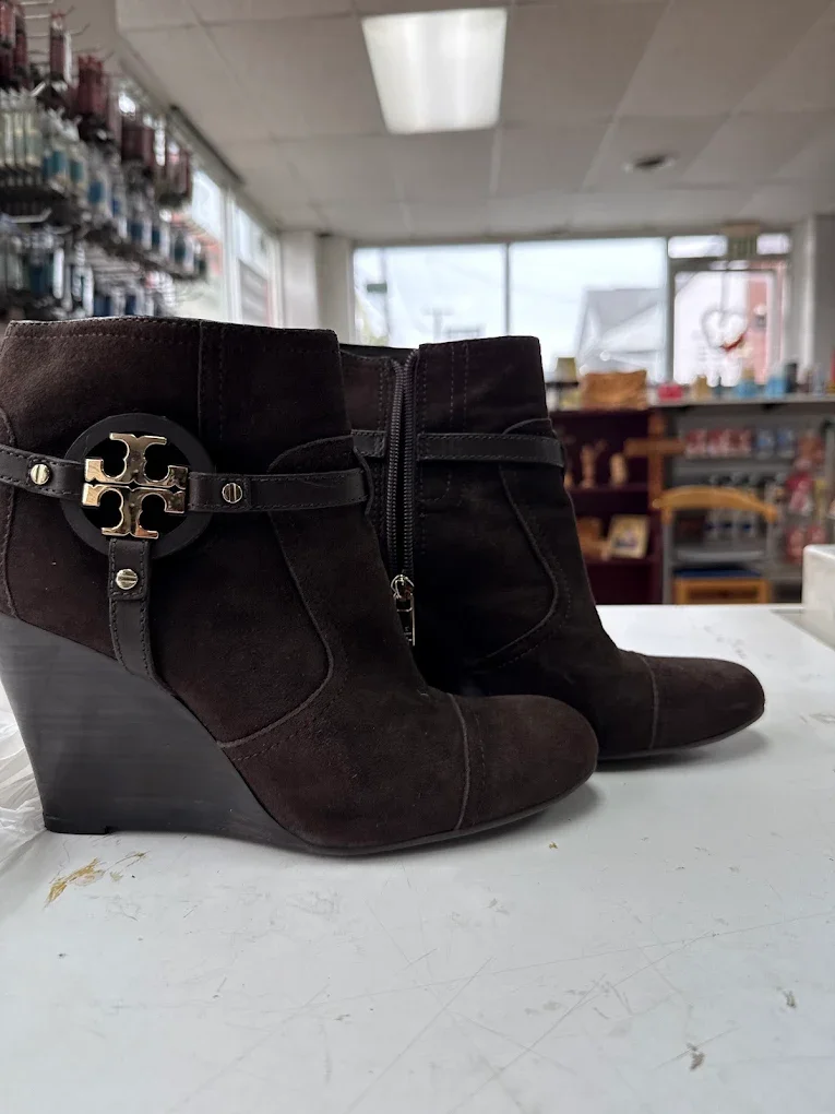 Black suede ankle boots with wedge heels and a decorative buckle strap, placed on a white surface in a store.