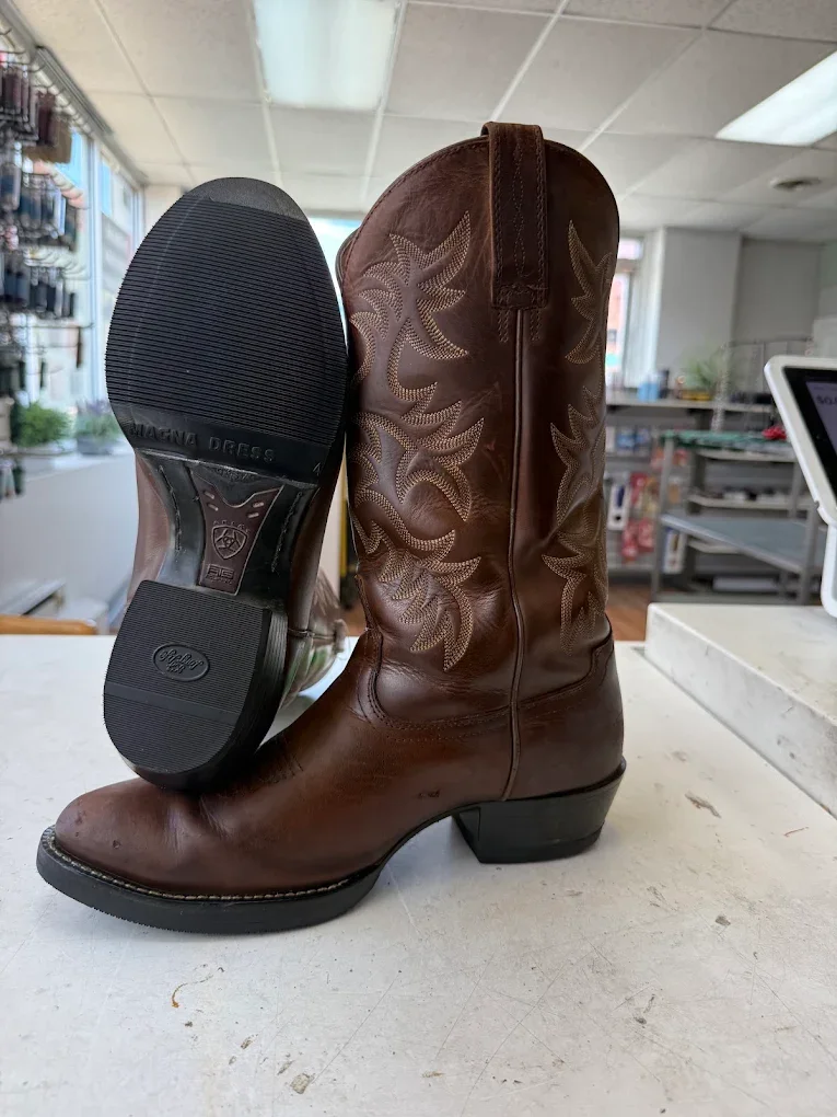 A pair of brown leather cowboy boots, one boot standing upright and the other tilted to show the sole, on a white surface in a store.
