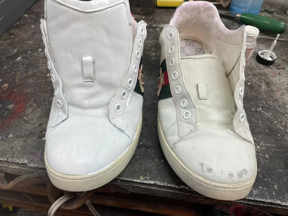 A pair of white high-top sneakers with a Gucci logo on the side, one of which has visible dirt and scuffs. The shoes are on a workbench with tools and supplies around.