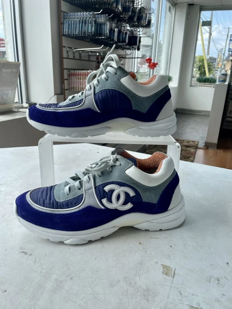 Pair of Chanel sneakers in blue, gray, and white with the Chanel logo on the side, displayed on a white surface inside a store.