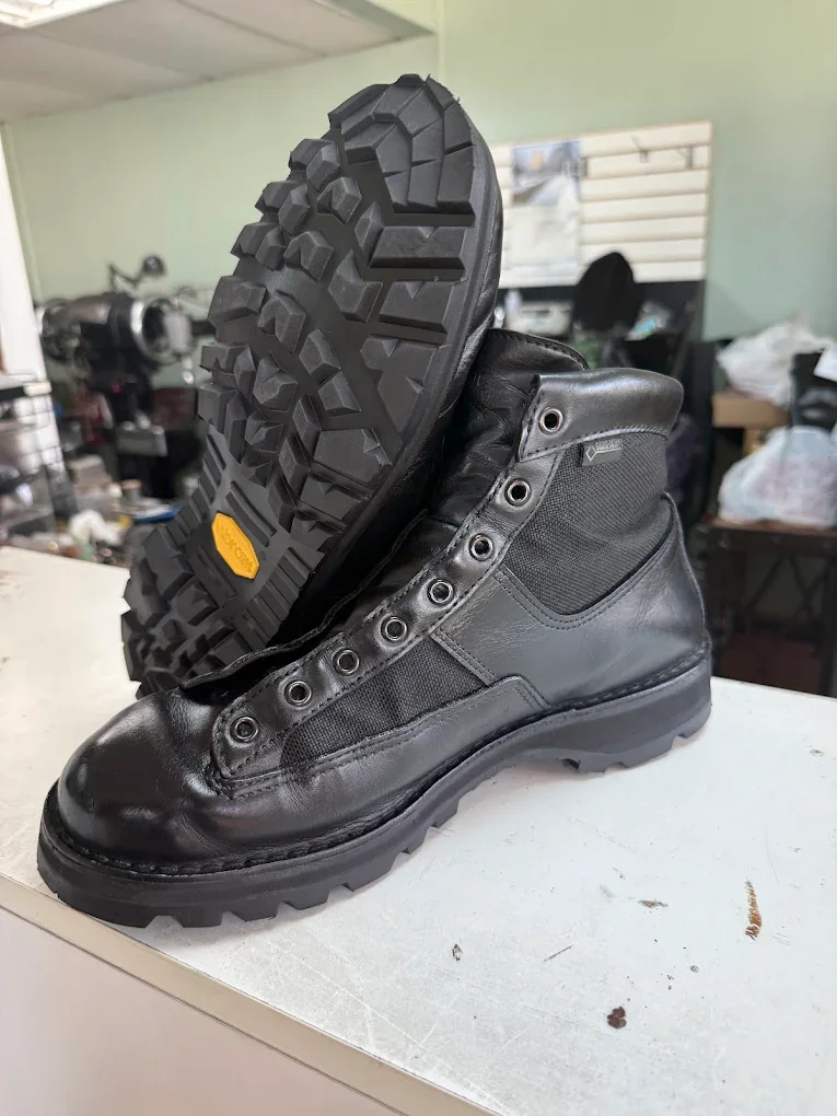 Black leather work boots with rugged soles, one boot standing upright and the other tilted to show the tread, on a white surface in a workshop setting.