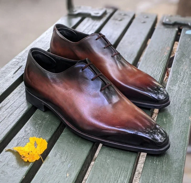 Pair of polished men's dress shoes with a brown to black gradient, placed on a green wooden bench with a yellow leaf nearby.