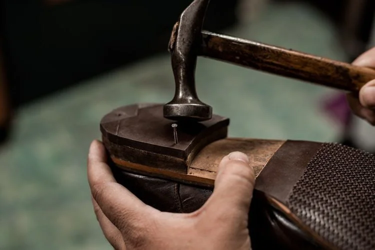 A person using a hammer and a nail to attach a black sole to a brown leather shoe in a workshop.