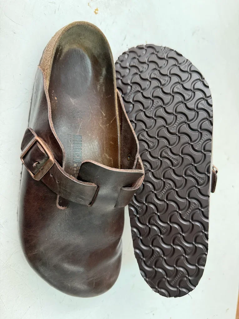 A worn brown leather Birkenstock sandal with an adjustable strap and buckle, placed next to a black rubber sole with a textured pattern.