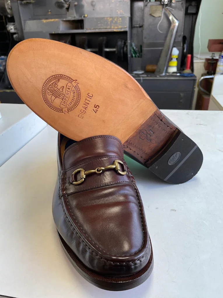 A pair of men's brown leather dress shoes, with one shoe lying on its side showing the sole that is labeled with size 45 and the brand 'Imperial Gigantic,' and the other shoe upright. The shoes are placed on a white surface in a workshop or shoe repair shop.