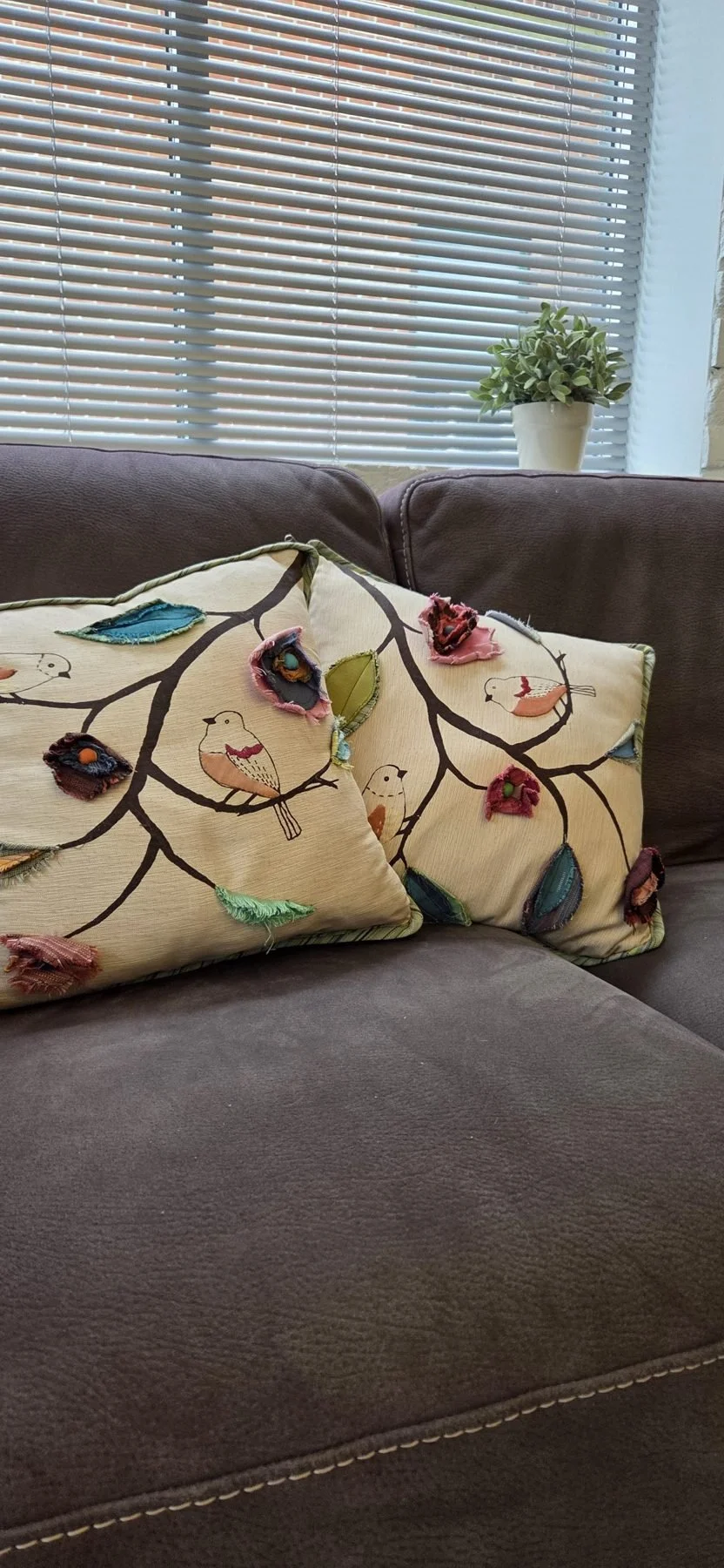 Living room with a brown knit sofa, decorative pillows with a bird and branch pattern, a window with closed blinds, and a potted plant on the window sill.