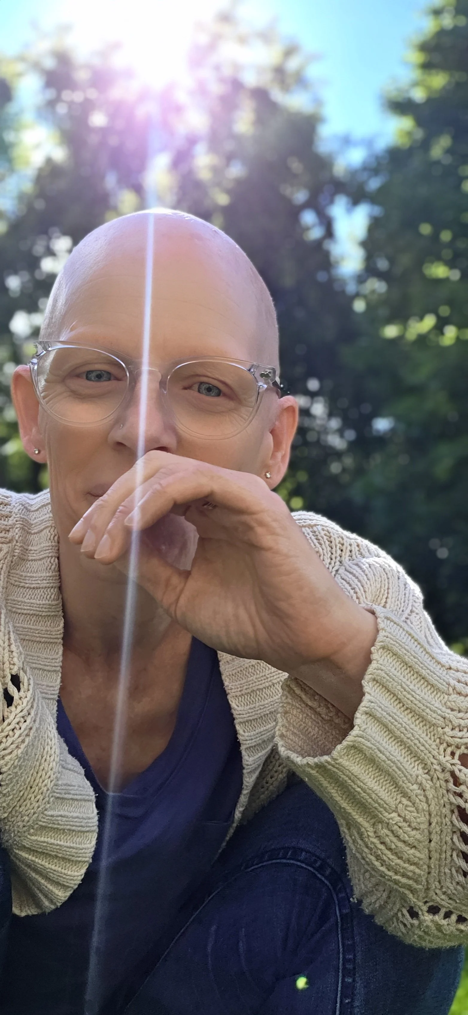 A woman with a bald head, glasses, and earrings, sitting outdoors with sunlight filtering through trees in the background, wearing a cream-colored knit cardigan over a dark top.