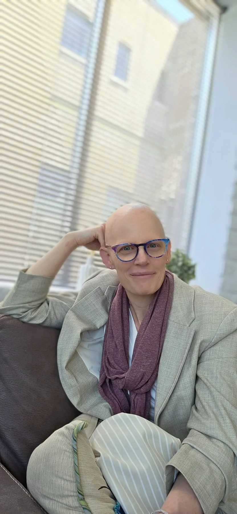 A person with a bald head, wearing colorful glasses, a beige blazer, a maroon scarf, and sitting on a sofa indoors near large windows.