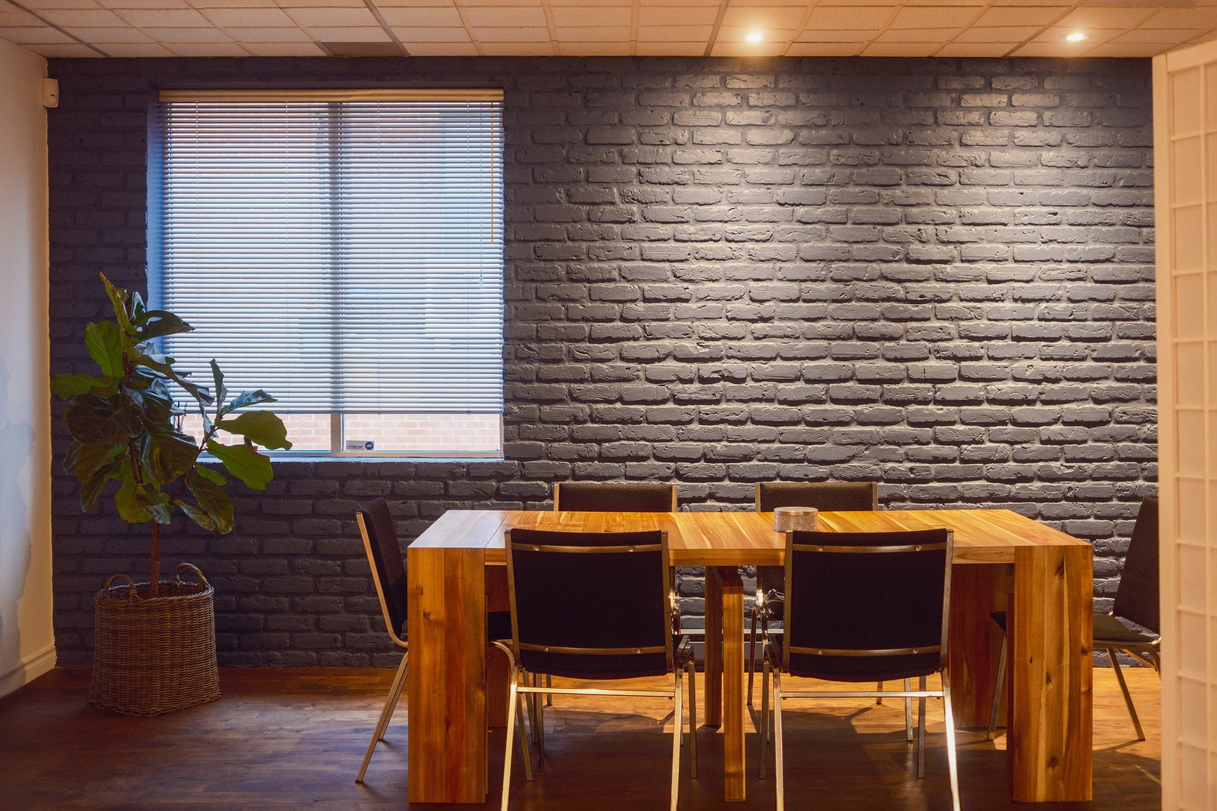 A dining area with a wooden table and black chairs in front of a dark gray brick wall. A window with blinds is on the wall, and a potted plant is in the corner next to a woven basket. Recessed ceiling lights illuminate the room.