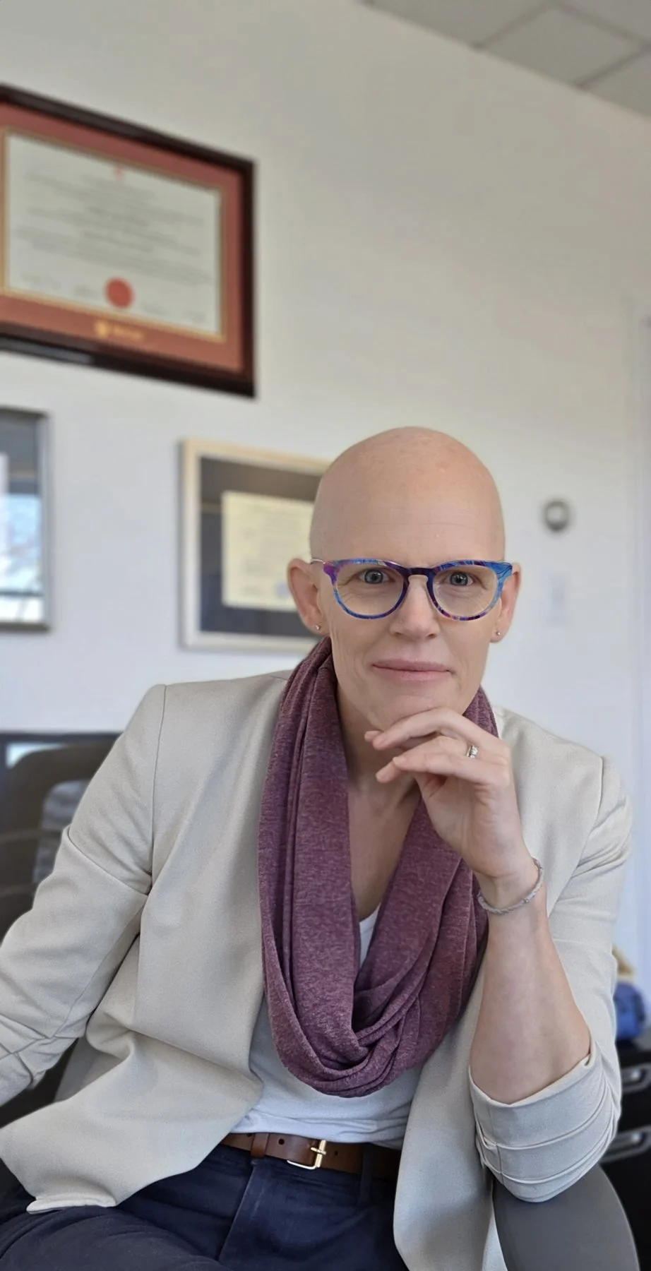 A woman with a bald head wearing colorful glasses and a beige blazer, sitting at a desk in an office with framed certificates on the wall behind her.