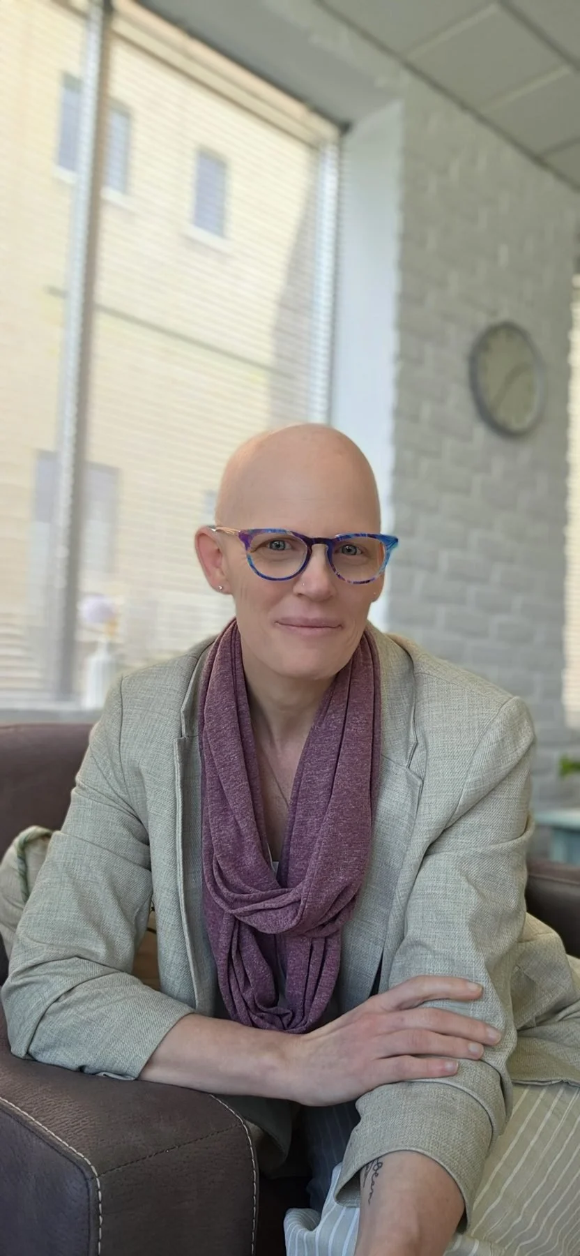 A woman with a bald head, wearing colorful glasses and a beige blazer with a purple scarf, sitting at a table in a well-lit indoor space with large windows and brick walls.
