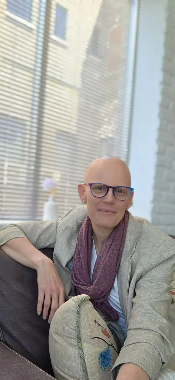 A woman with glasses and a shaved head sitting on a couch near a window with blinds.
