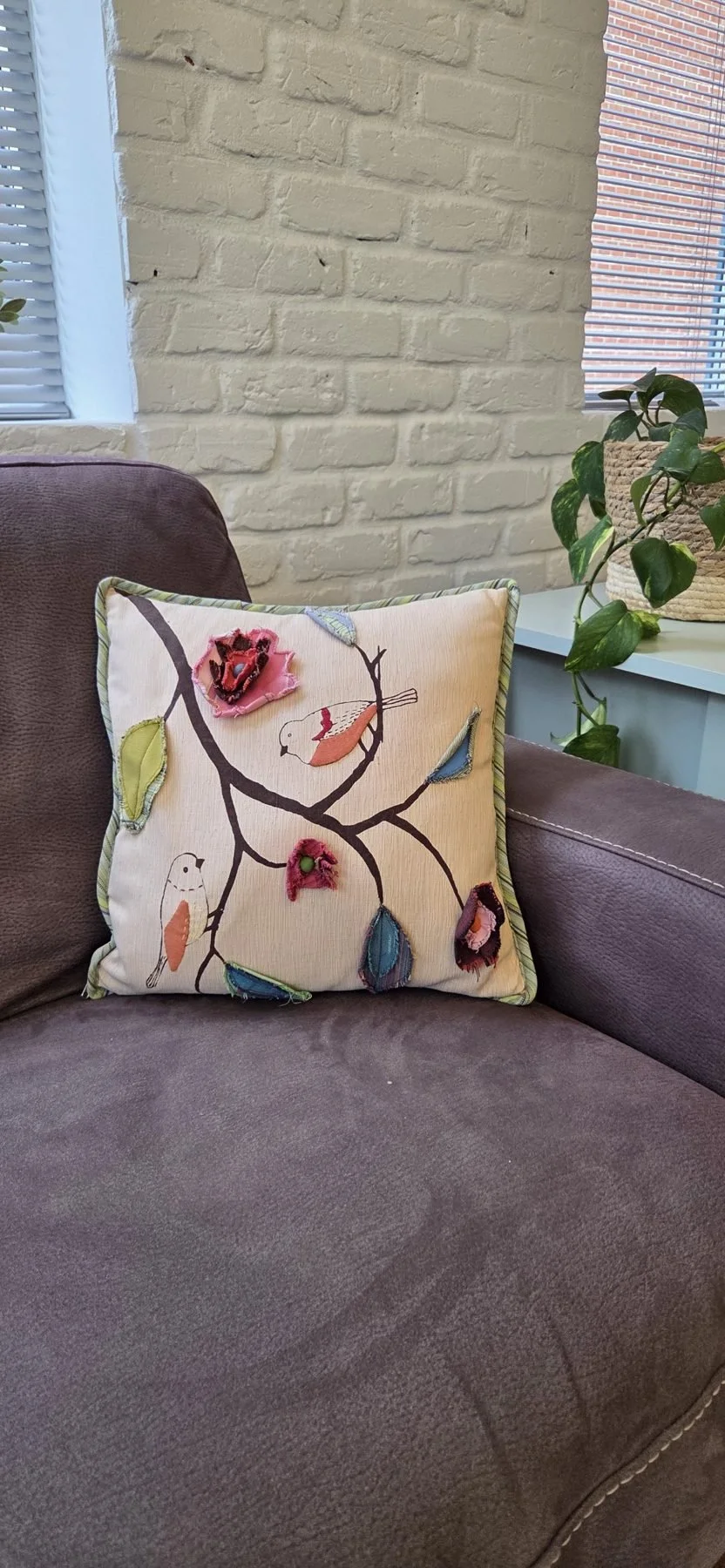 Decorative pillow with embroidered bird and branch design on a brown sofa in a room with brick wall and potted plant nearby.