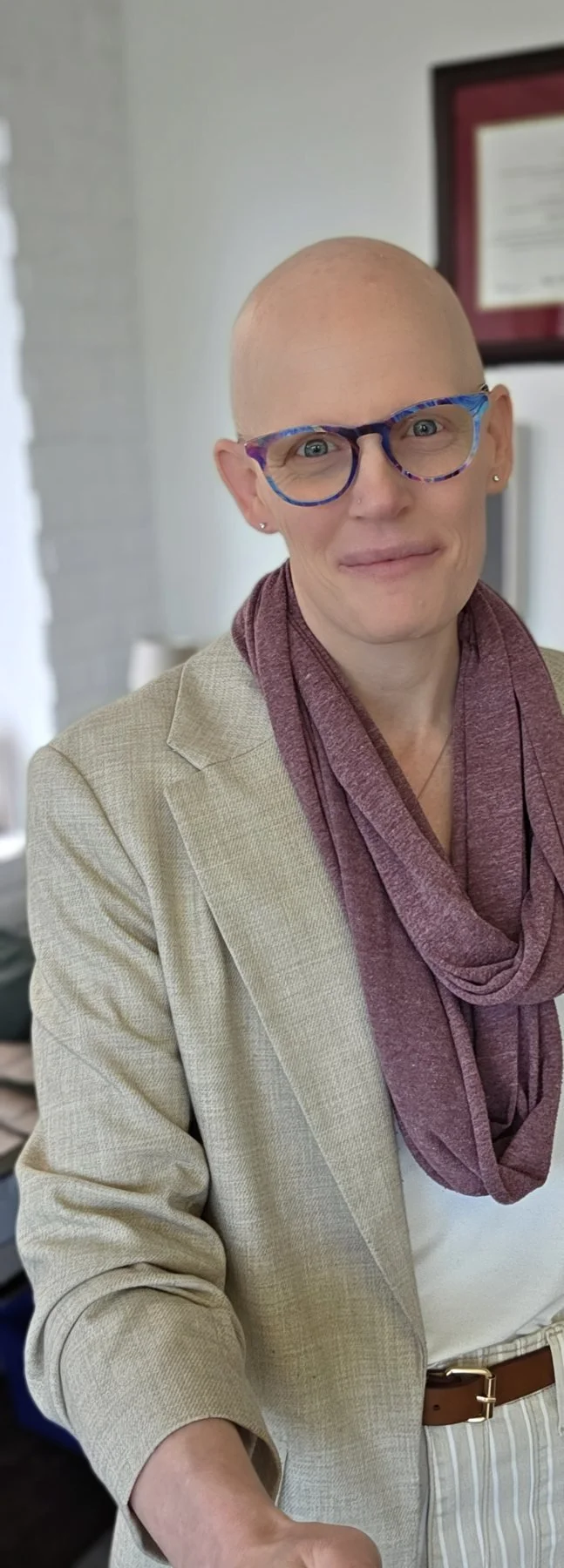 A woman with a bald head wearing colorful glasses, a beige blazer, a white top, and a maroon scarf, indoors with a framed certificate in the background.