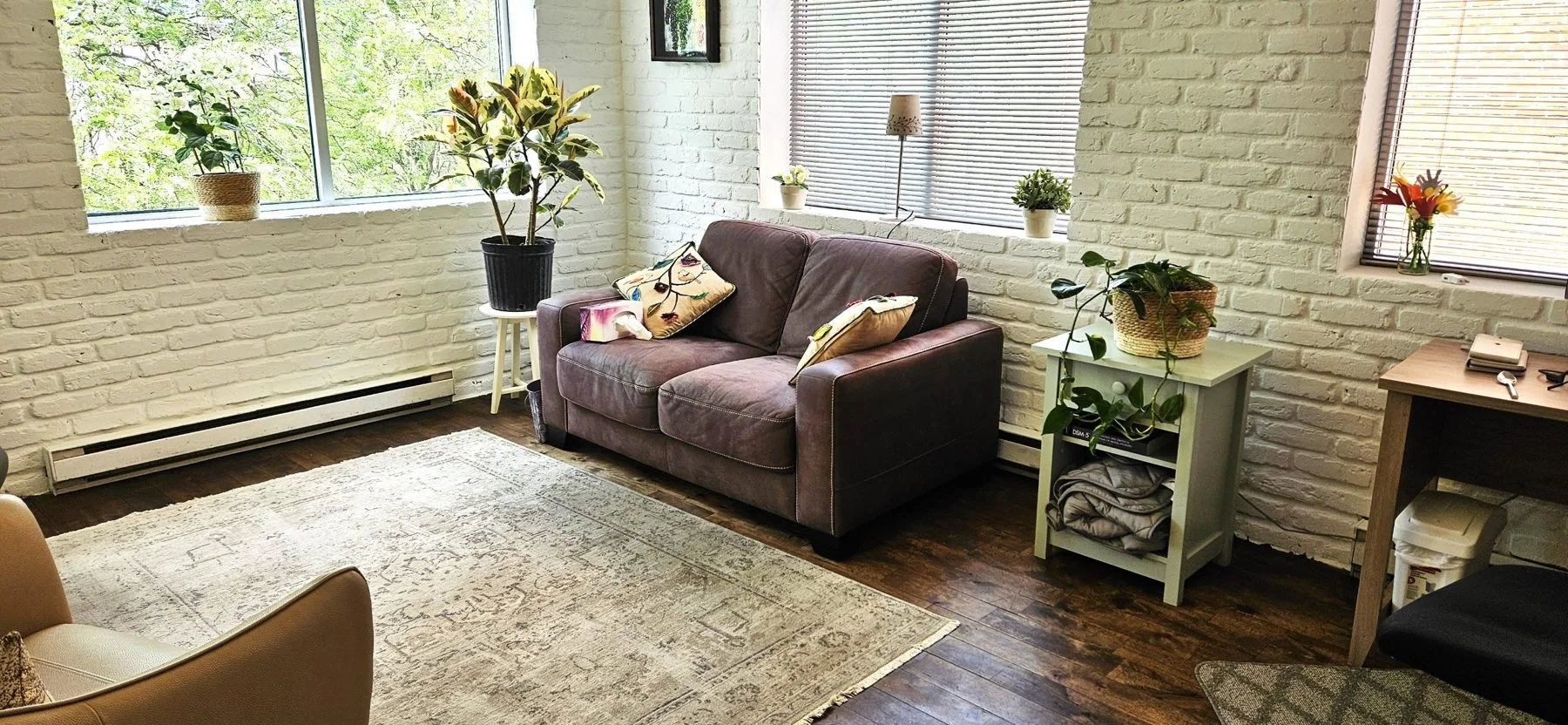 Living room with white brick walls, large windows, a brown leather loveseat with decorative pillows, a beige area rug, and various potted plants on white and wooden tables.