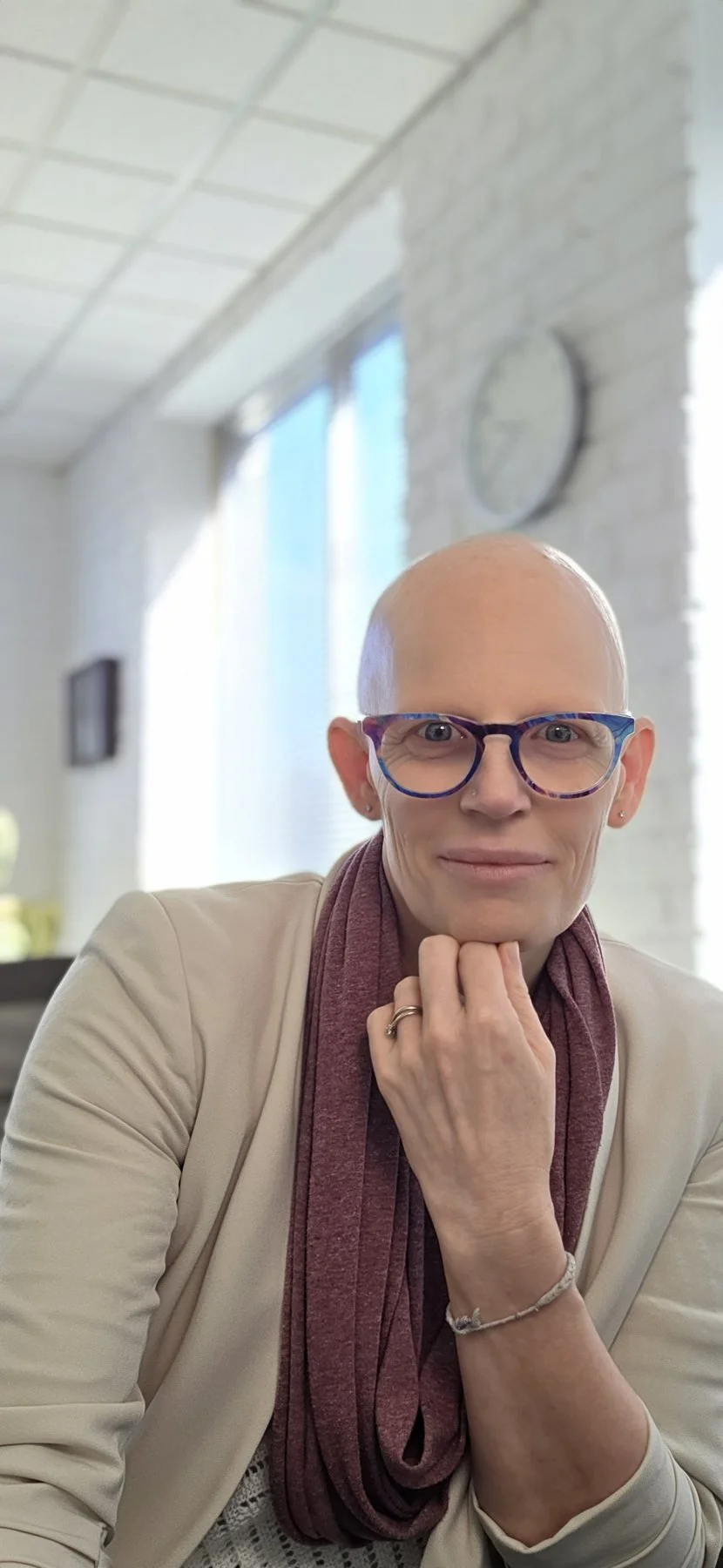 A woman with a bald head, wearing glasses, a beige blazer, and a maroon scarf, sitting indoors in front of a large window with sunlight, holding her chin with her left hand.