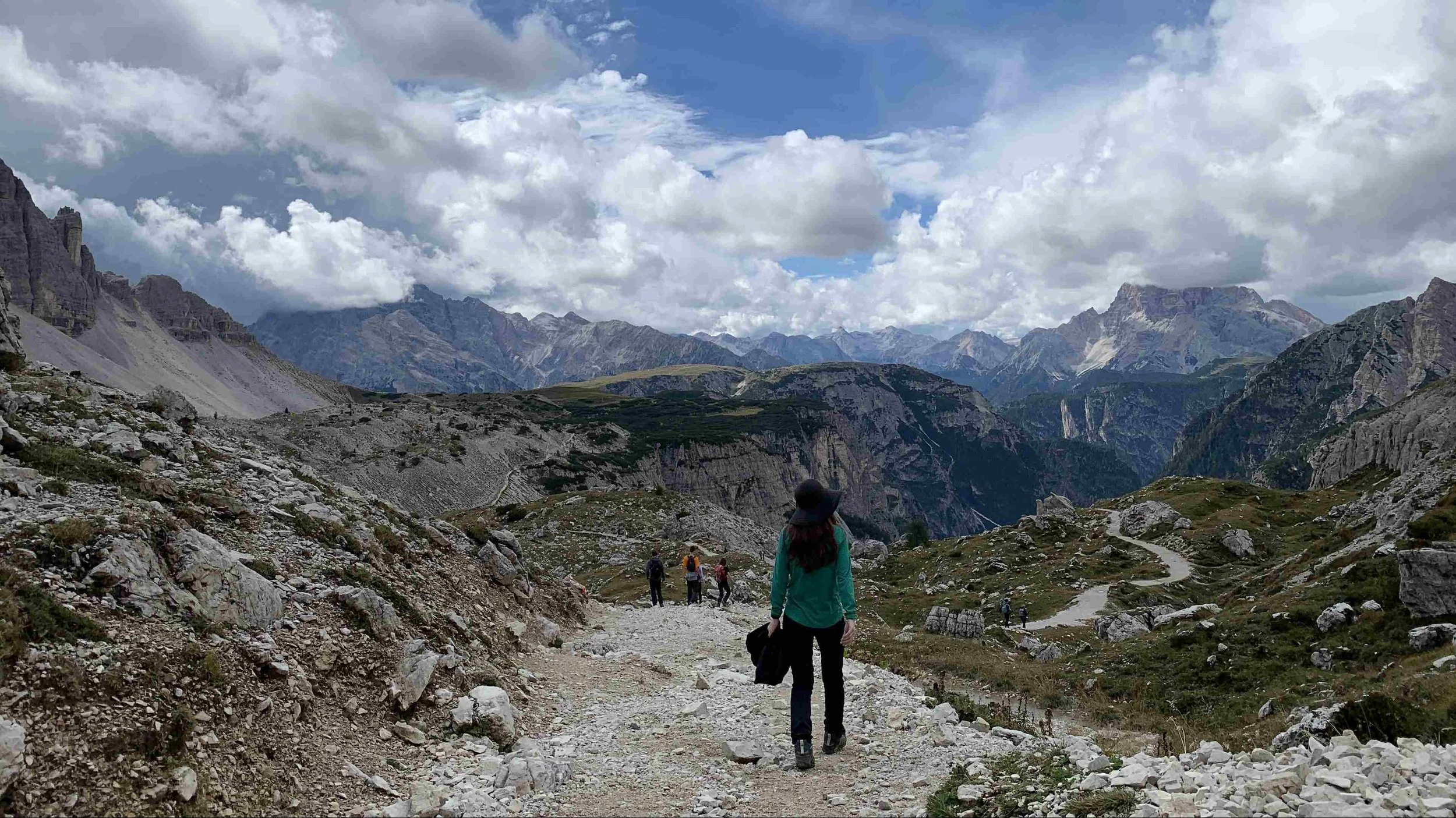 Hikers walking on a rocky trail in a mountainous landscape with clouds and peaks in the distance.