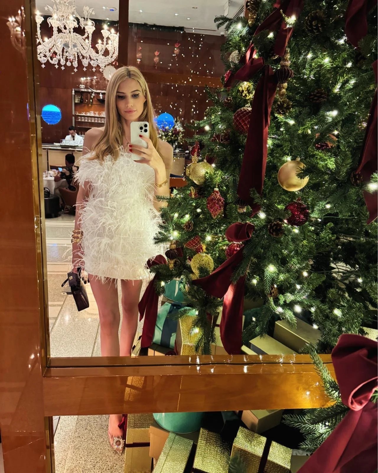 Sienna Blakely, a VIP London escort and luxury dinner companion, taking a glamorous mirror selfie in a white feather mini dress beside a beautifully decorated Christmas tree.