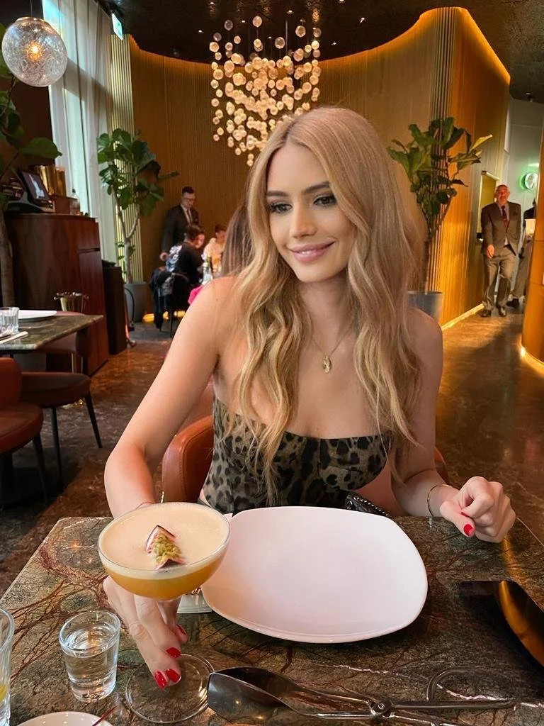 High-class London escort Sienna Blakely enjoying an elegant restaurant outing in a leopard-print dress, with long blonde waves and a cocktail at the table.