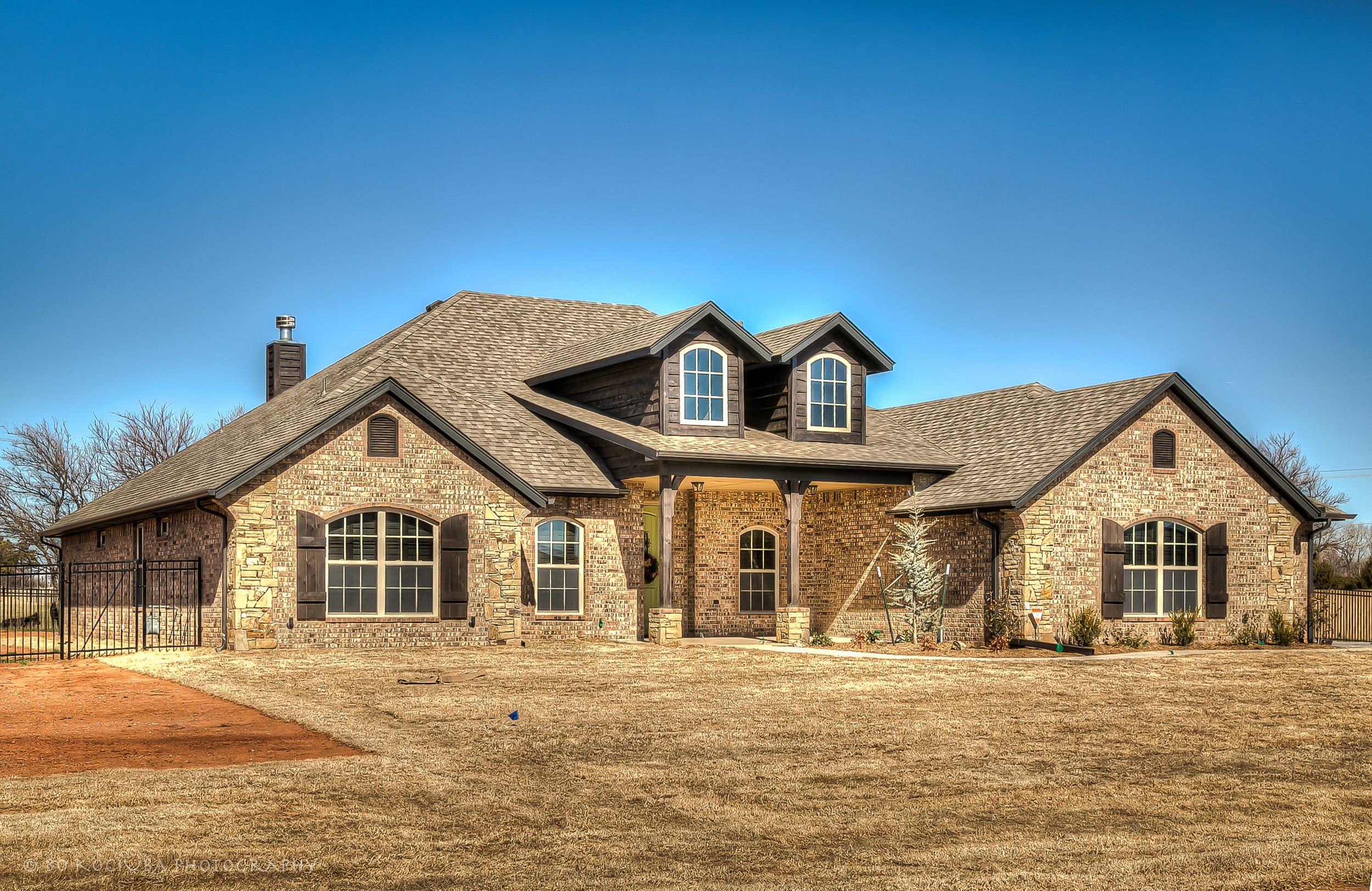 Large brick home in Chisholm Run, Yukon, Oklahoma, featuring multiple gabled rooflines, dormer windows, a covered porch, and a spacious front yard.