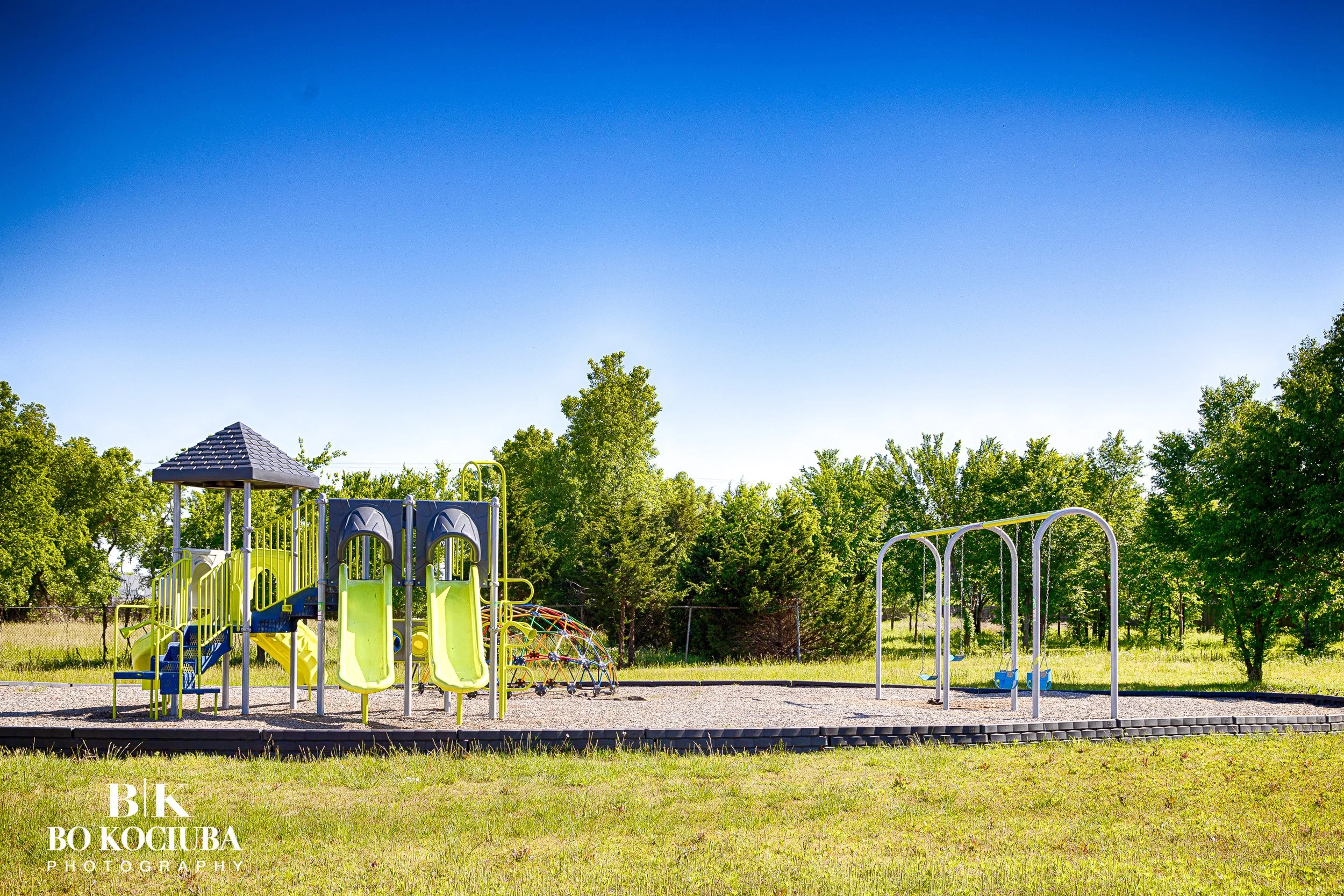 Colorful playground equipment including slides, climbing structures, and rings set in a grassy park with trees and a clear blue sky.