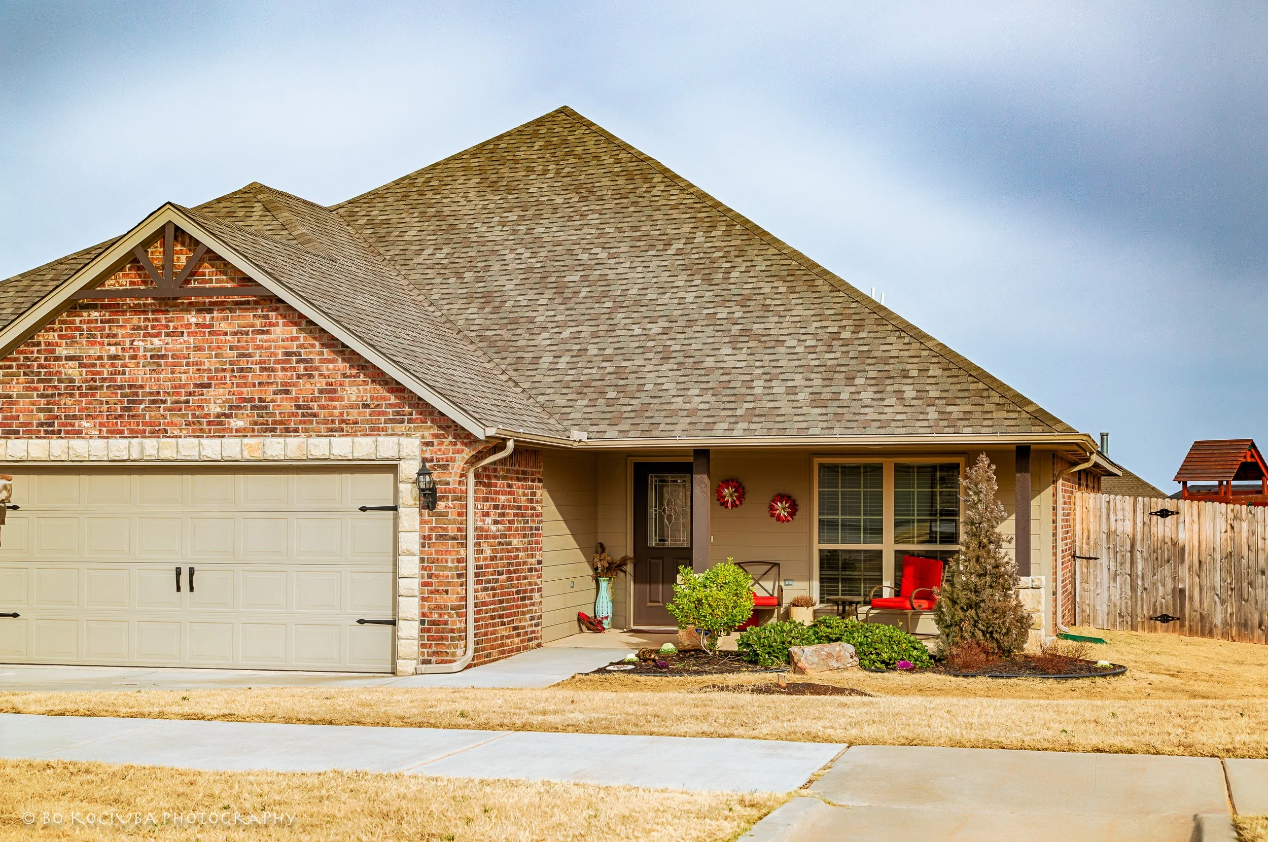 Brick home in Chisholm Crossing, Yukon, Oklahoma, featuring an attached garage and covered front porch decorated with seating, potted plants, and seasonal wreaths.