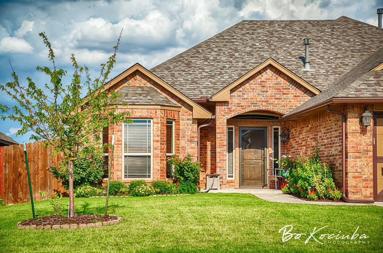 Single-story brick home in Chisholm Crossing, Yukon, Oklahoma, featuring an arched covered entry, landscaped shrubs, a young tree, and a green front lawn.