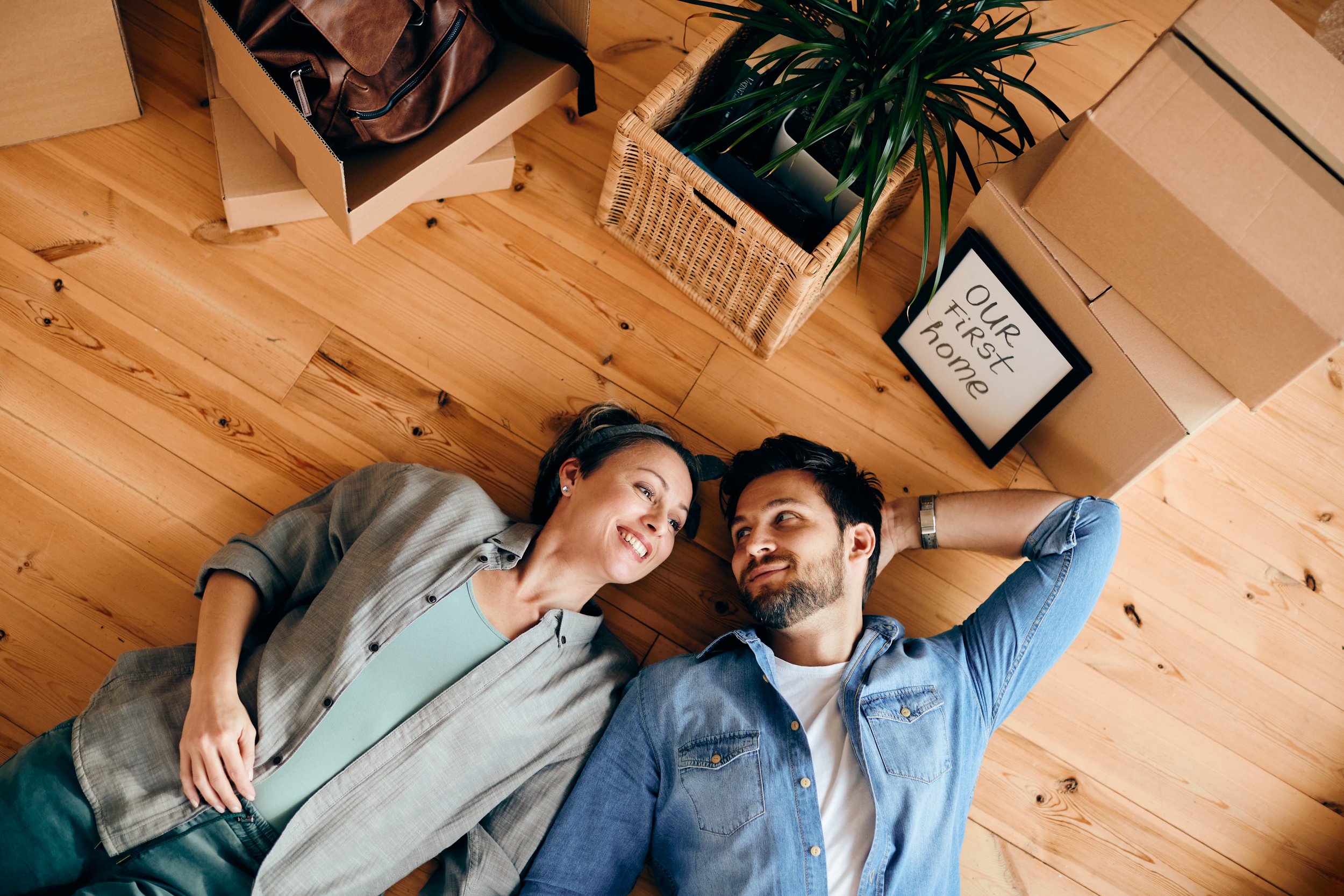 Two new homeowners relaxing on the floor of their first home, surrounded by moving boxes and a framed “Our First Home” sign.