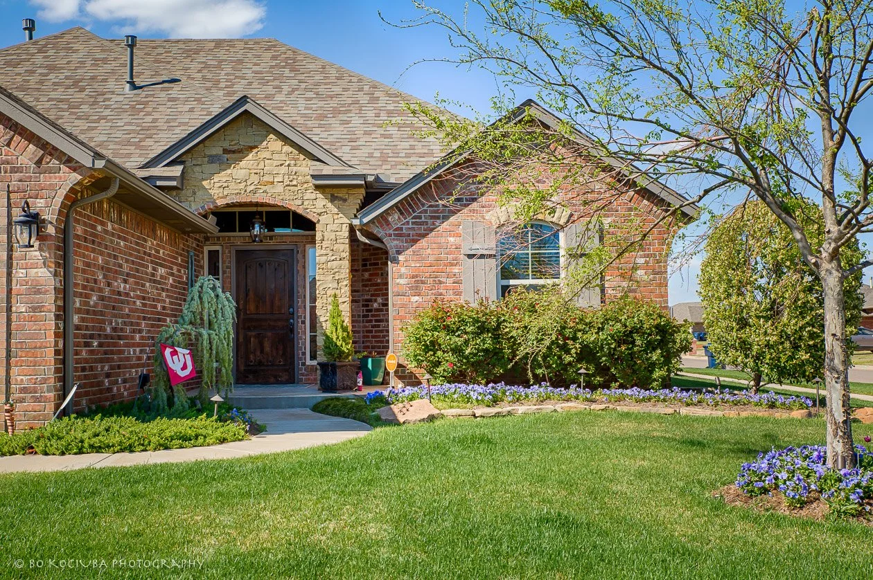 Brick and stone home in Sabram Estates, Yukon, Oklahoma, with a covered front entry, flowering shrubs, mature trees, and a landscaped lawn.