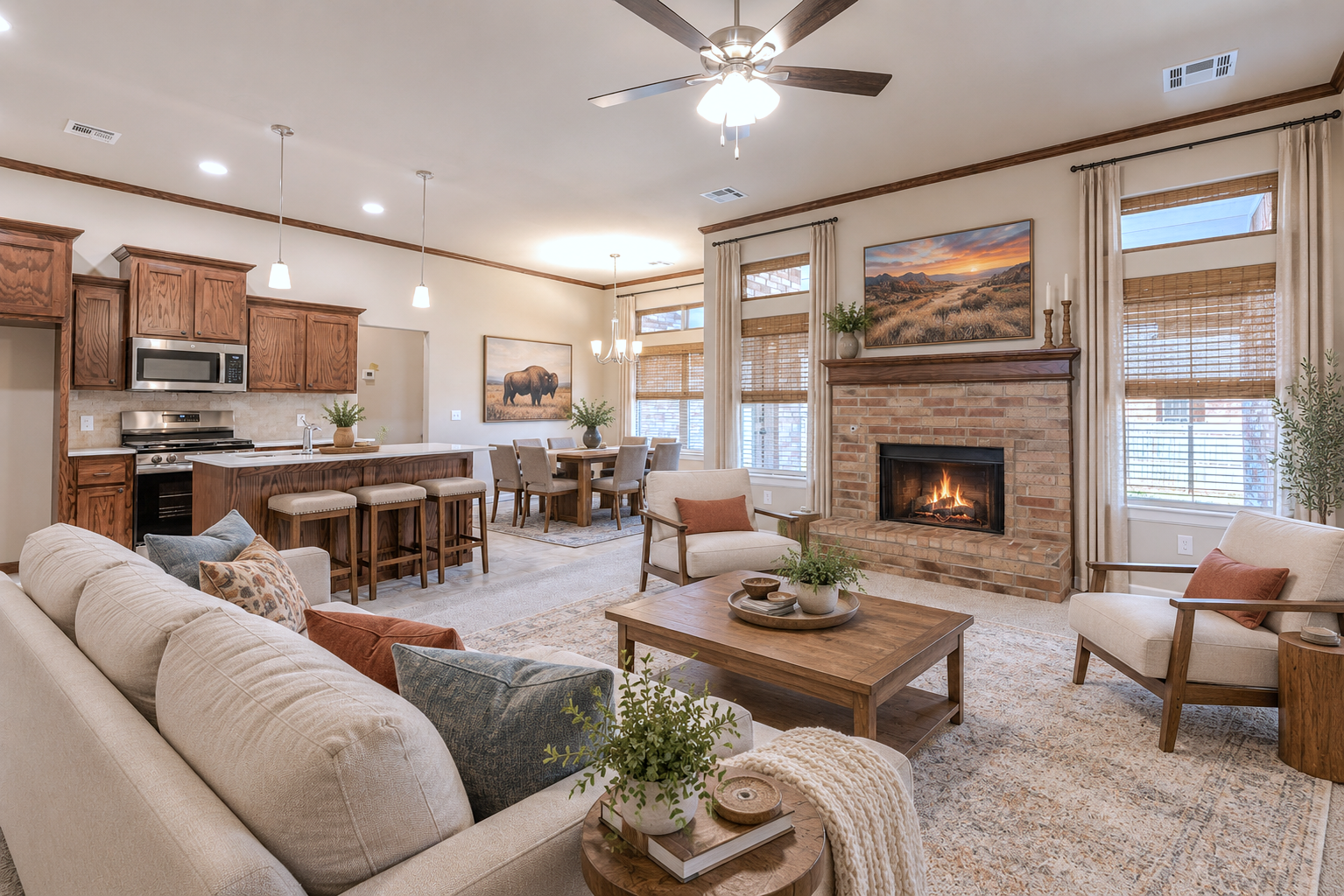 A spacious, cozy living room with a brick fireplace, beige sofas and armchairs, a wooden coffee table with decorative plants, large windows with blinds and curtains, a dining area with a chandelier, and a kitchen with wooden cabinets and an island with bar stools.