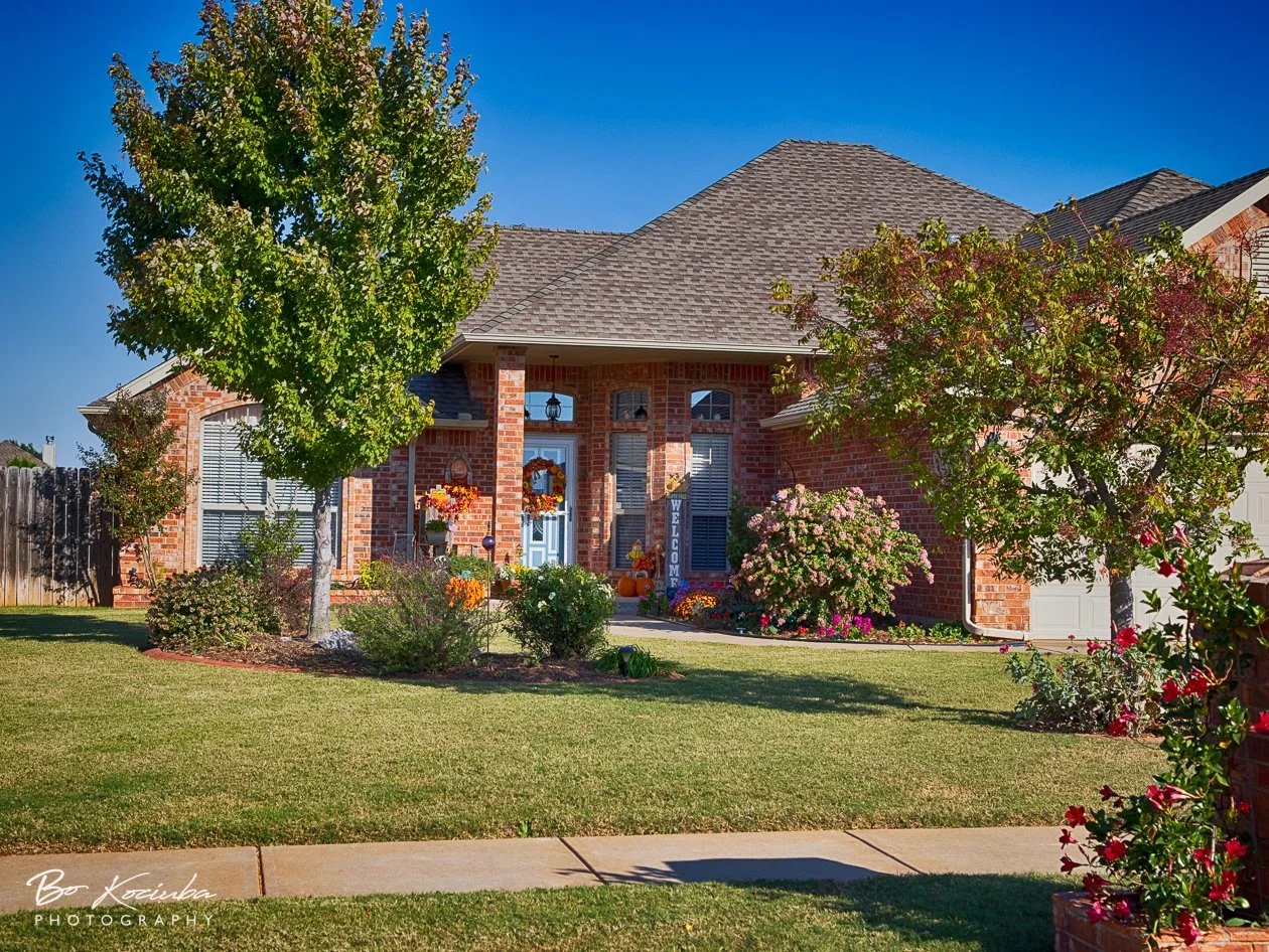 Brick home in Hunters Hill, Mustang, Oklahoma, featuring a covered front porch with autumn decorations, landscaped flower beds, and a well-kept lawn.