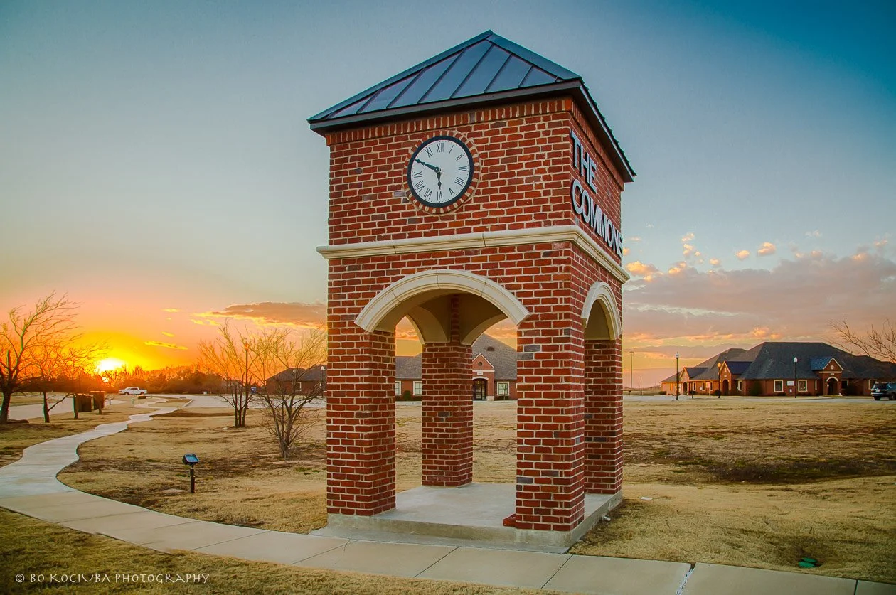 Freestanding brick clock tower beside a sidewalk, photographed at sunset with open land and commercial development in the background in Yukon, OK.